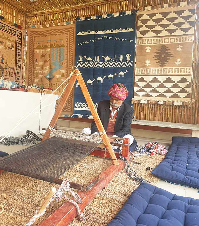 A weaver at a traditional loom with woven rugs hanging behind him